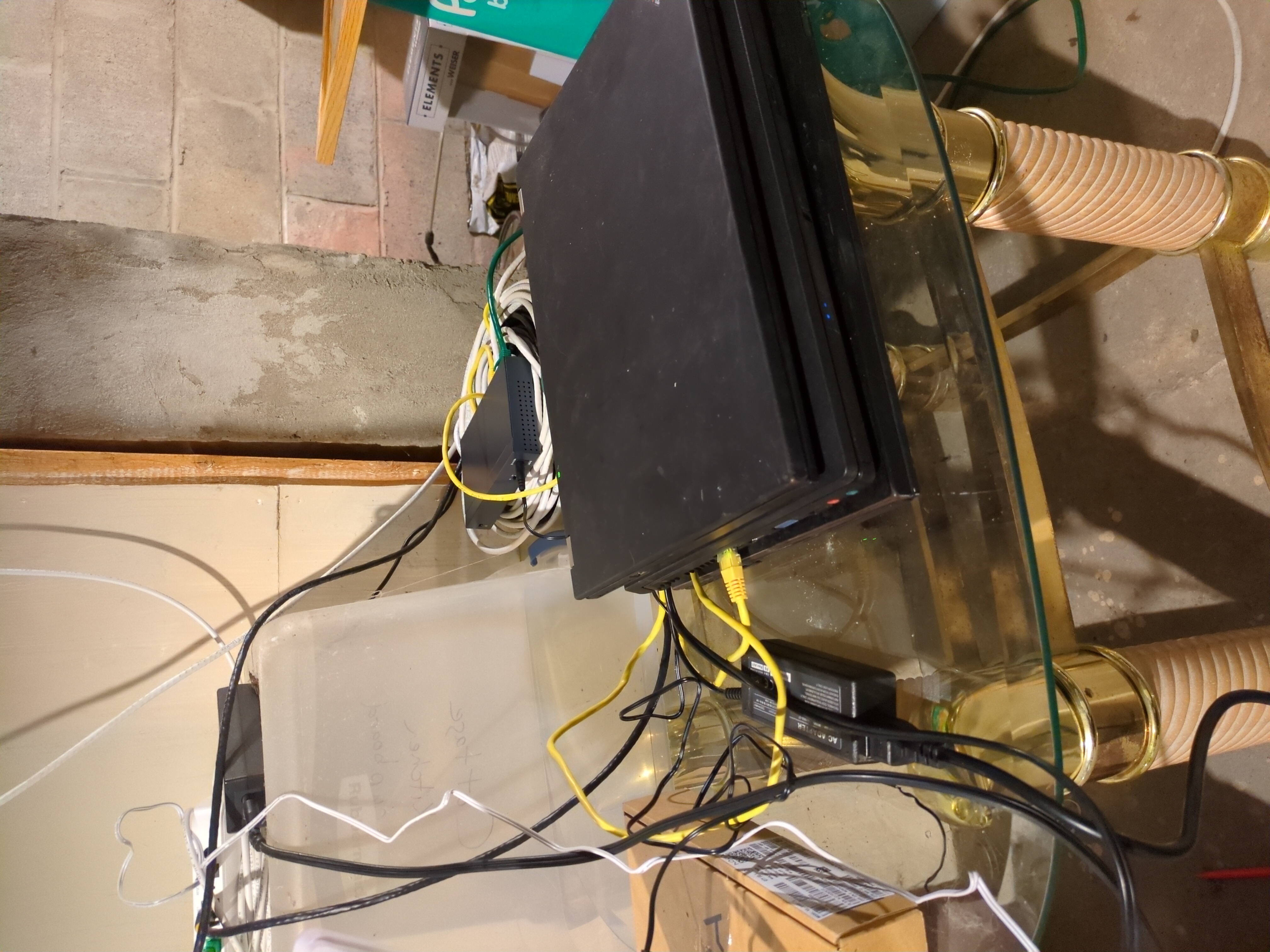 A pile of three laptops, a switch and a fibre modem connected by ethernet cables, sitting on a table in an unfinished basement.